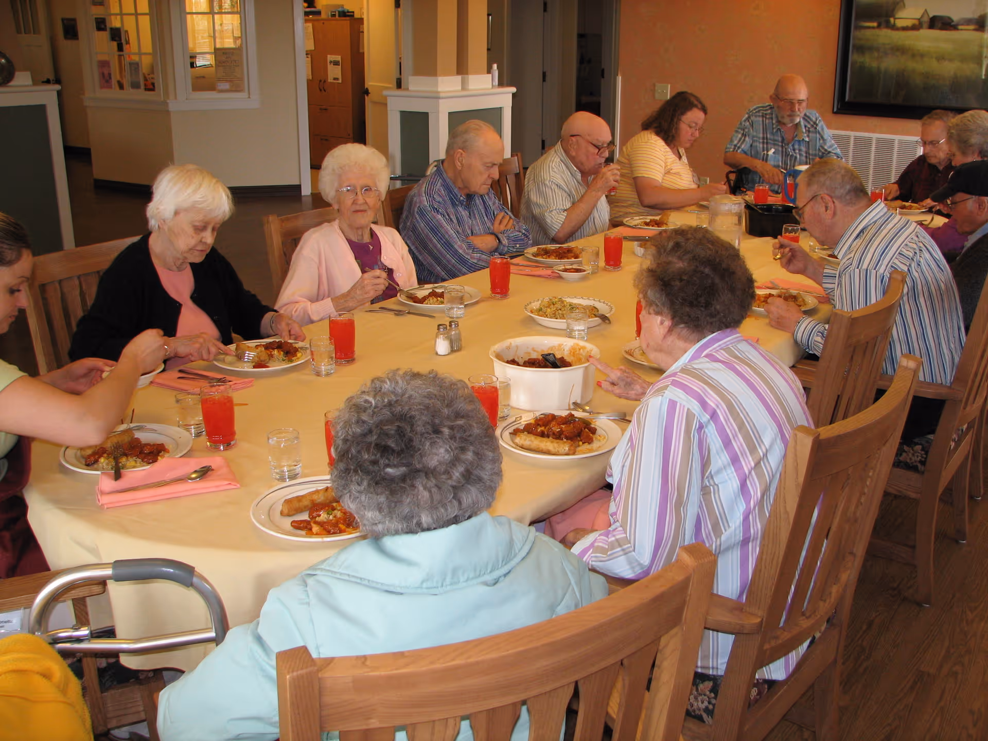 A group of elderly people sitting around a large dining table enjoying a meal together in a well-lit room. The table is covered with a yellow tablecloth and has plates of food, glasses of water, and red drinks. The room has wooden chairs and a warm, inviting atmosphere.