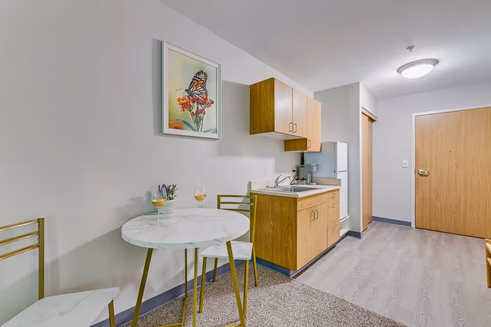 A small kitchenette area in a senior living facility featuring wooden cabinets, a white countertop with a sink, and a white refrigerator. Adjacent to the kitchenette is a round white marble table with two matching chairs, each with a glass of white wine on the table. A framed picture of a butterfly on flowers hangs on the wall above the table. The floor is a combination of carpet and light wood laminate, and a wooden door is visible in the background.