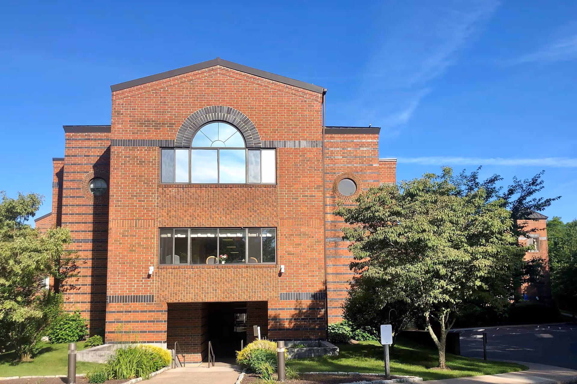 Exterior view of a multi-story brick building with large windows and an arched window at the top center. The building is surrounded by green trees and bushes under a clear blue sky.