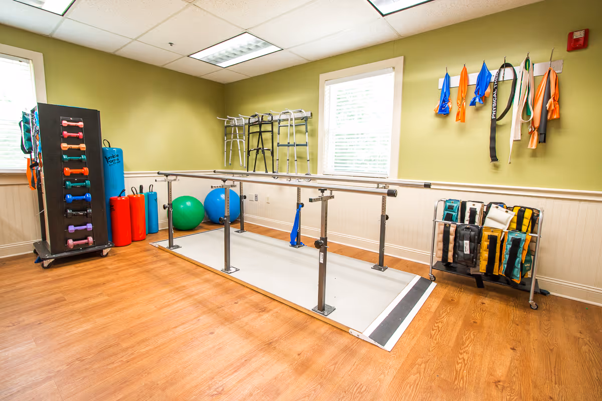 A bright rehabilitation exercise room with parallel bars, resistance bands, dumbbells, exercise balls and walkers.