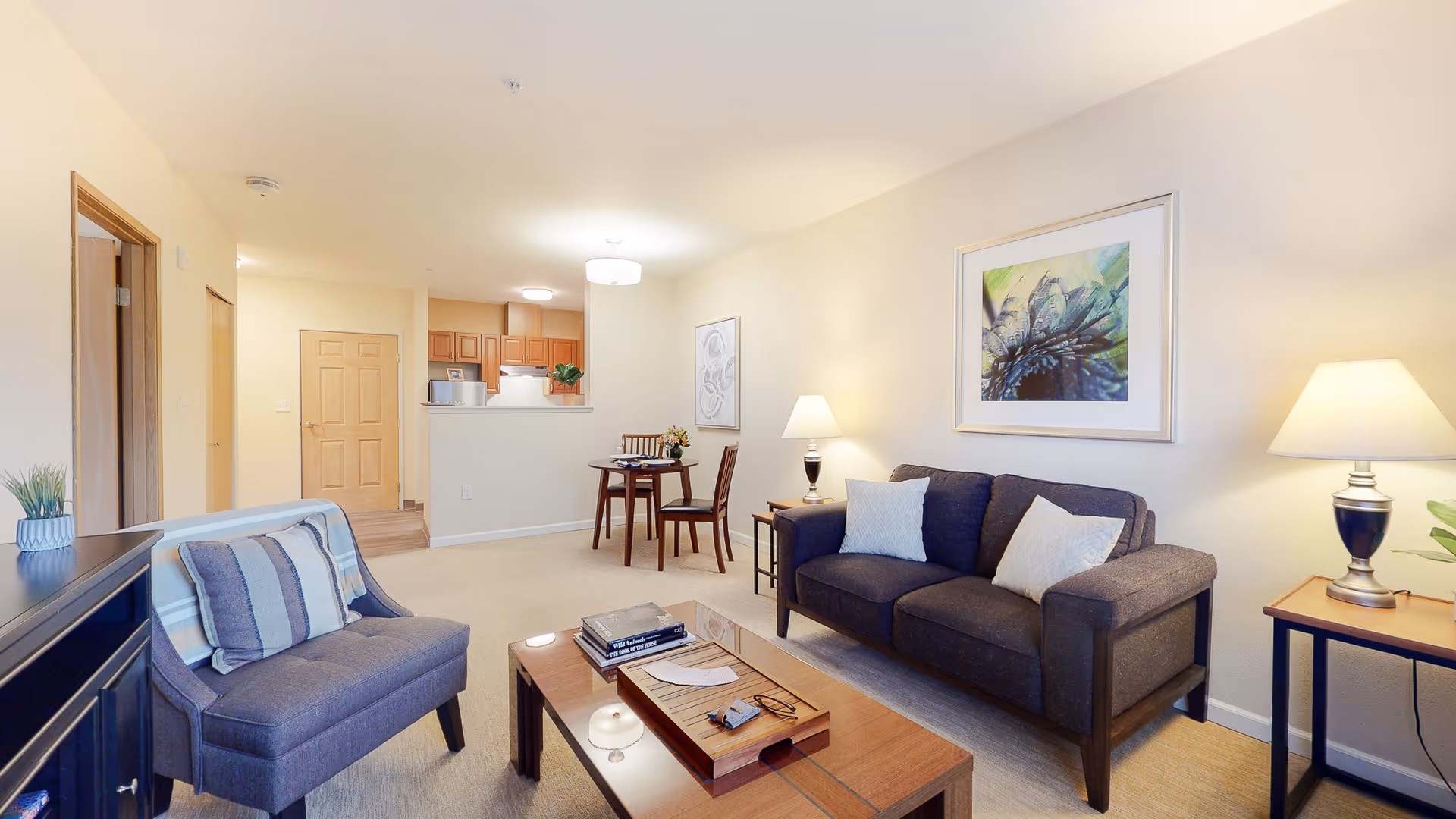 Bright furnished living room with a sofa, armchair, coffee table, and a small dining area with a view into the kitchen.