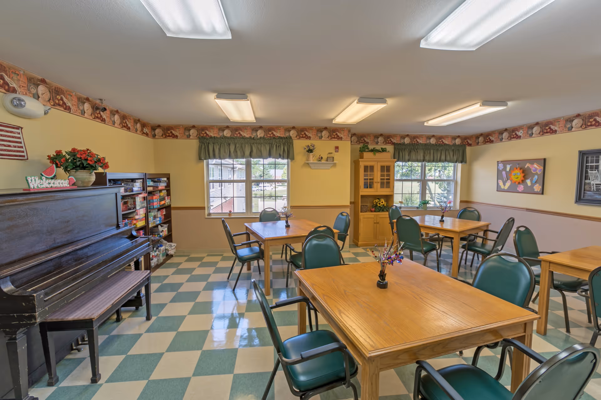 A well-lit common room with several wooden tables and green cushioned chairs arranged neatly. The room has a checkered green and white floor, yellow walls with a decorative border near the ceiling, and two windows with green valances letting in natural light. There is a black upright piano with a bench on the left side, a shelf filled with board games, and a wooden cabinet with decorative items. The room is decorated with flowers, a small American flag, and colorful artwork on the walls.