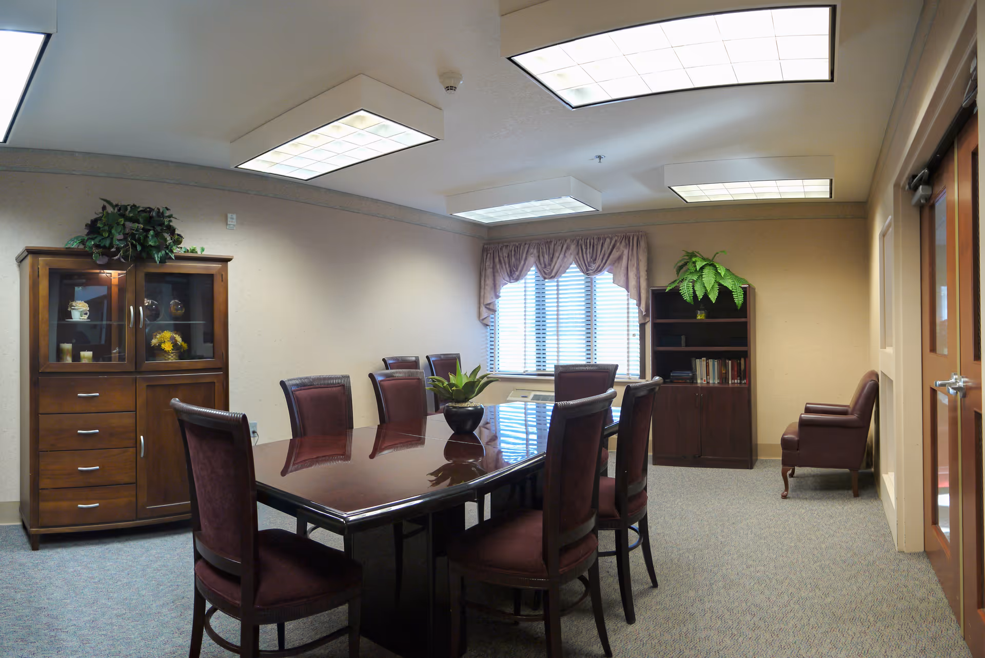 A conference or meeting room with a large polished wooden table surrounded by eight upholstered chairs. The room has beige walls, a window with blinds and a valance, a wooden cabinet with glass doors and decorative items, a bookshelf with books and a potted plant, and a single armchair near the door. The ceiling has multiple rectangular fluorescent light fixtures.