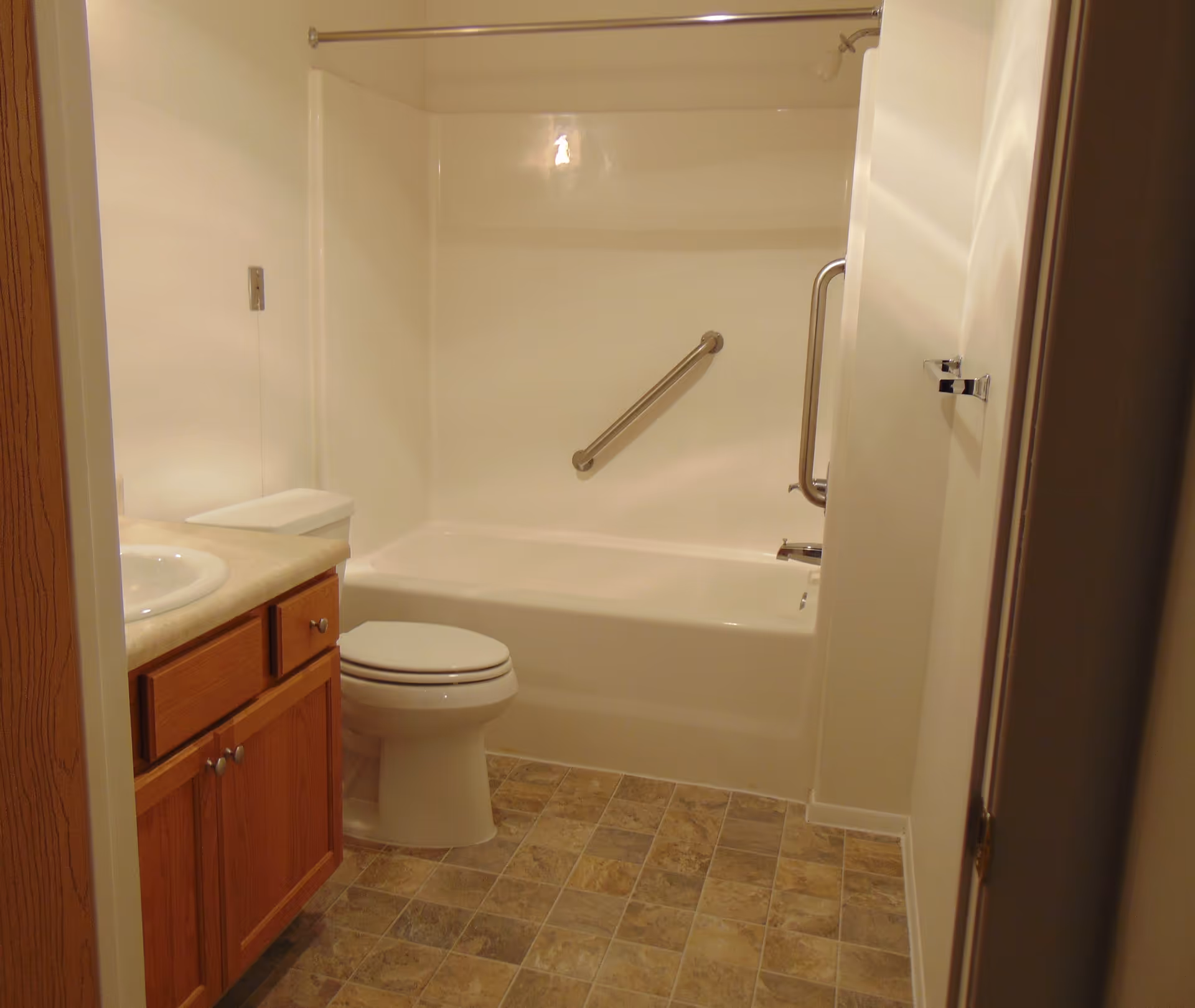 A clean bathroom featuring a white bathtub with two metal grab bars, a white toilet, a wooden vanity with a beige countertop and a white sink, and tiled flooring in shades of brown and beige.