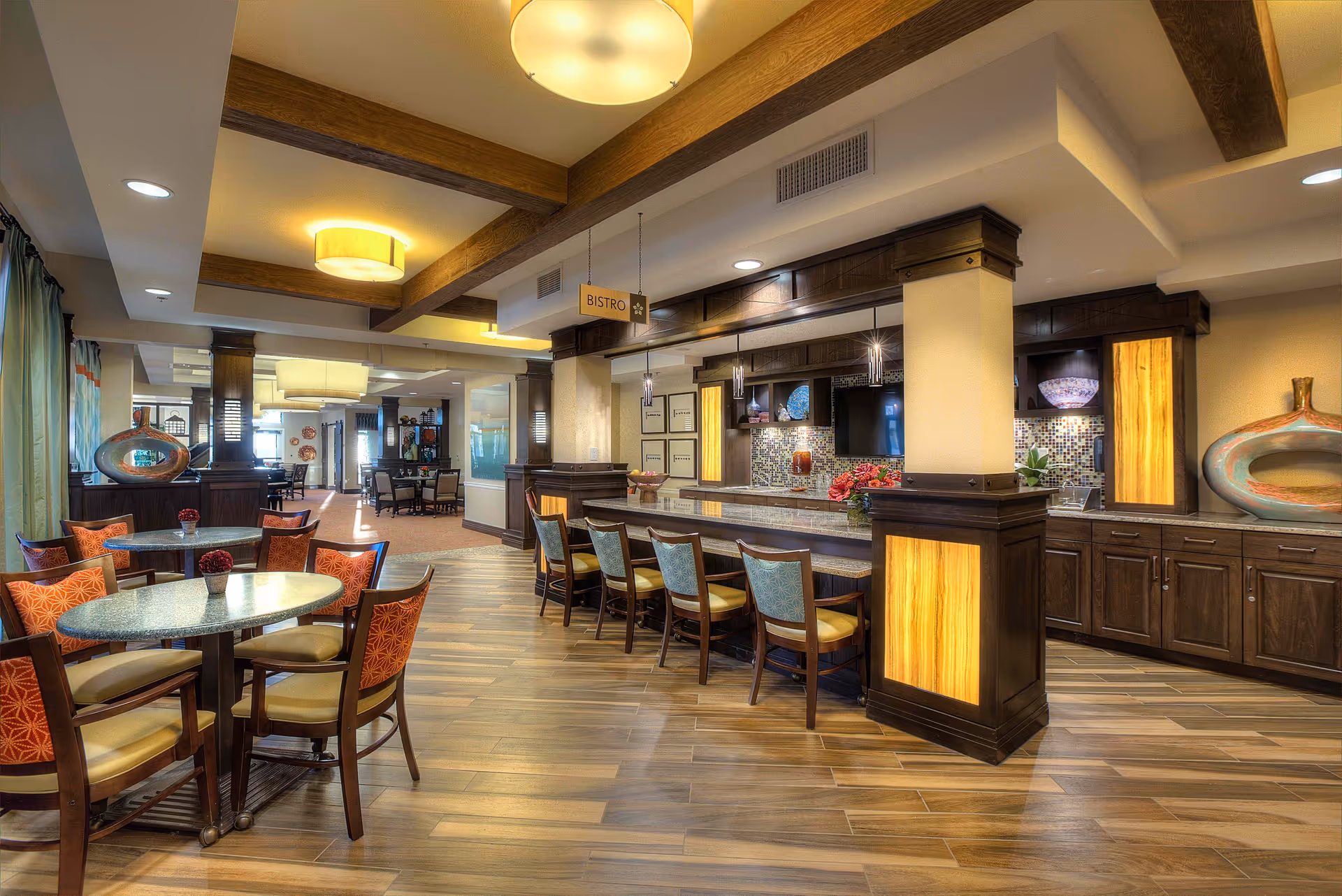 Interior view of a bright and spacious bistro area in MorningStar Assisted Living & Memory Care of Albuquerque featuring round tables with orange cushioned chairs, a long counter with high chairs, warm wood flooring, decorative lighting, and modern cabinetry with accent lighting.
