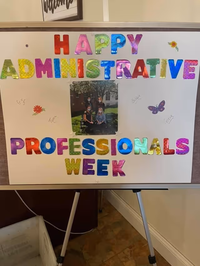 A colorful poster on an easel with the words 'HAPPY ADMINISTRATIVE PROFESSIONALS WEEK' spelled out in large, shiny, multicolored letters. The poster includes a small photo of four people wearing masks sitting and standing outside near a tree. There are small decorative stickers of flowers and a butterfly around the text and photo.