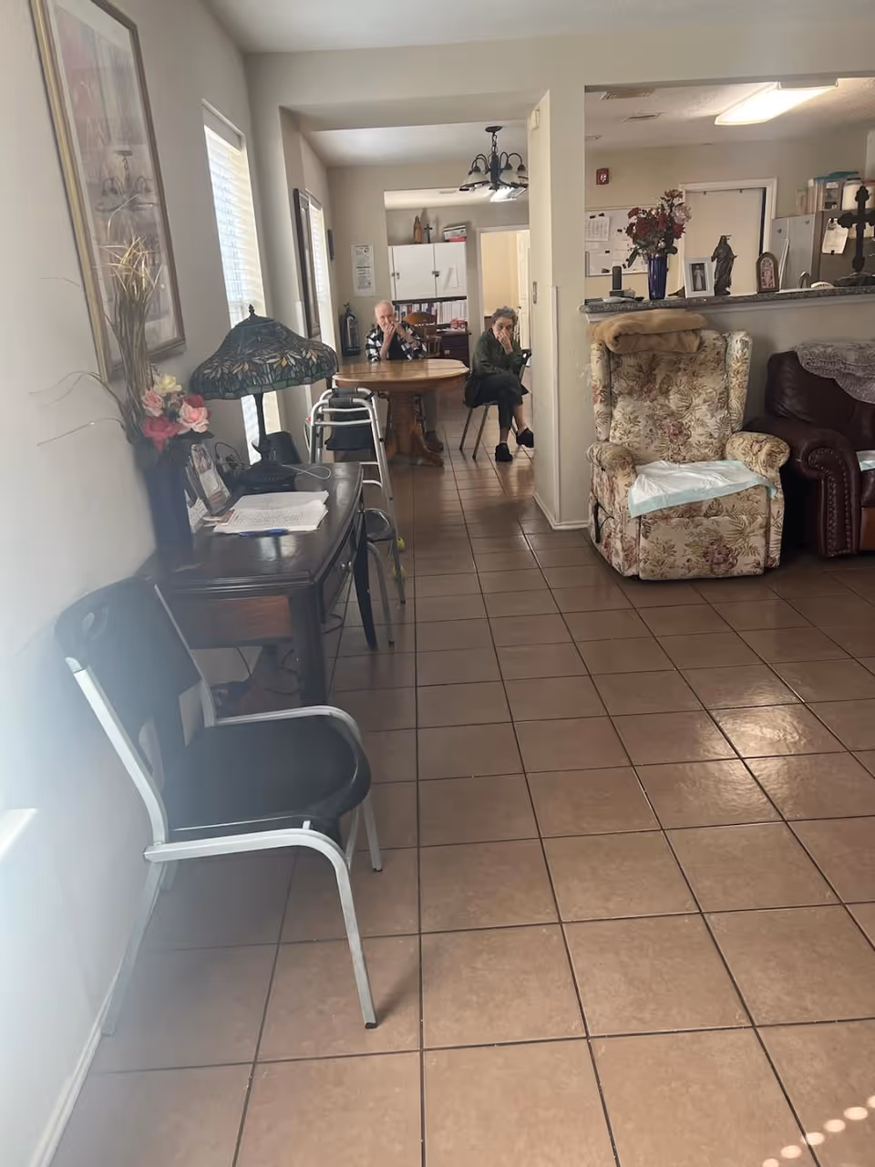 Interior view of a senior living facility showing a tiled floor hallway with chairs along the left wall and a wooden table with two elderly people sitting at it in the background. On the right side, there is a floral patterned armchair with a blanket on the backrest and a leather couch partially visible. The area appears to be a common living space with a kitchen visible in the background.