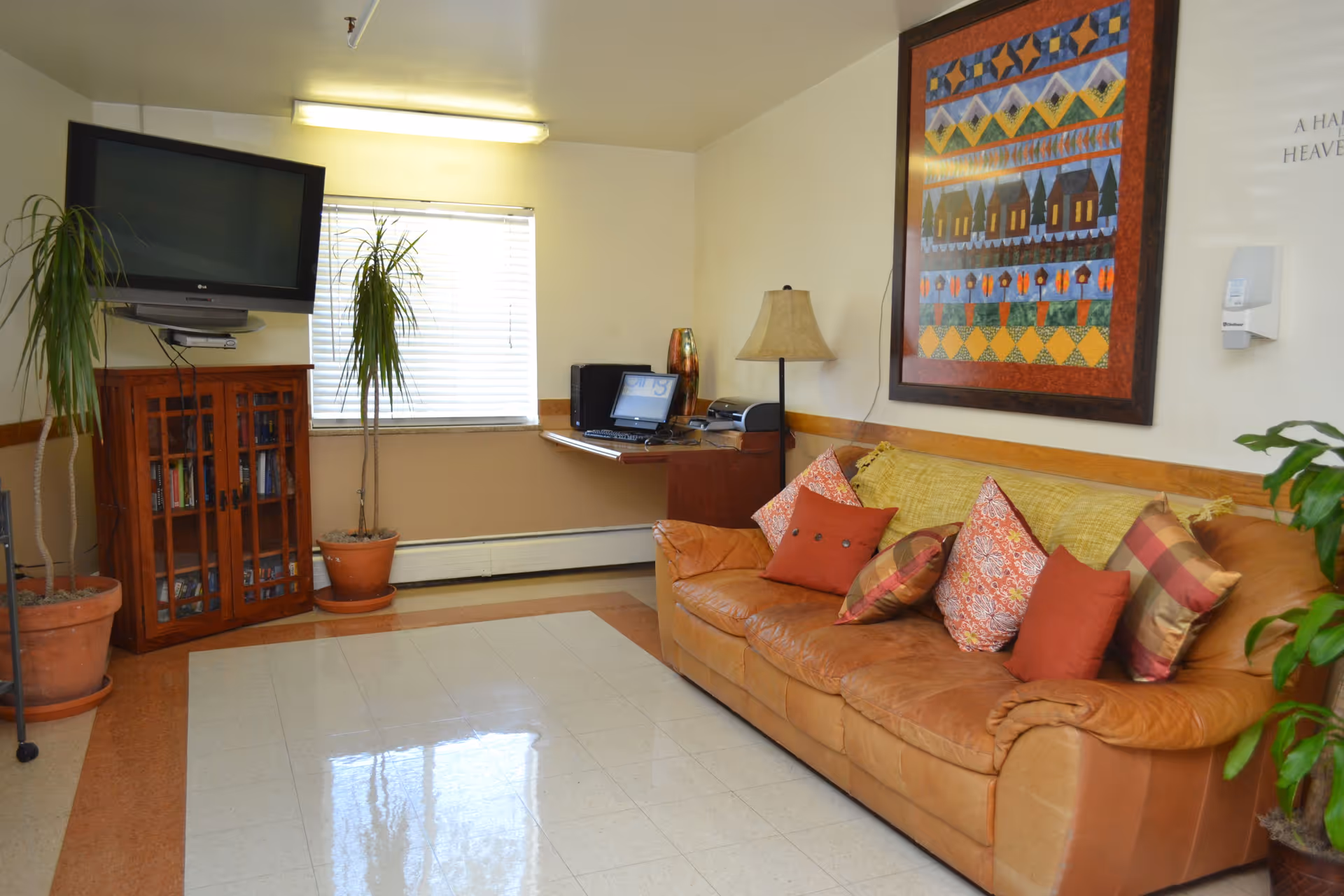 A cozy living room area with a brown leather couch adorned with multiple colorful pillows. Above the couch hangs a framed quilt-like artwork. To the left, there is a wooden cabinet with glass doors, a flat-screen TV mounted on the wall above it, and two potted plants. A window with blinds is behind the plants. A small desk with a laptop and printer is in the corner next to a floor lamp. The floor has a tiled pattern with a brown border.