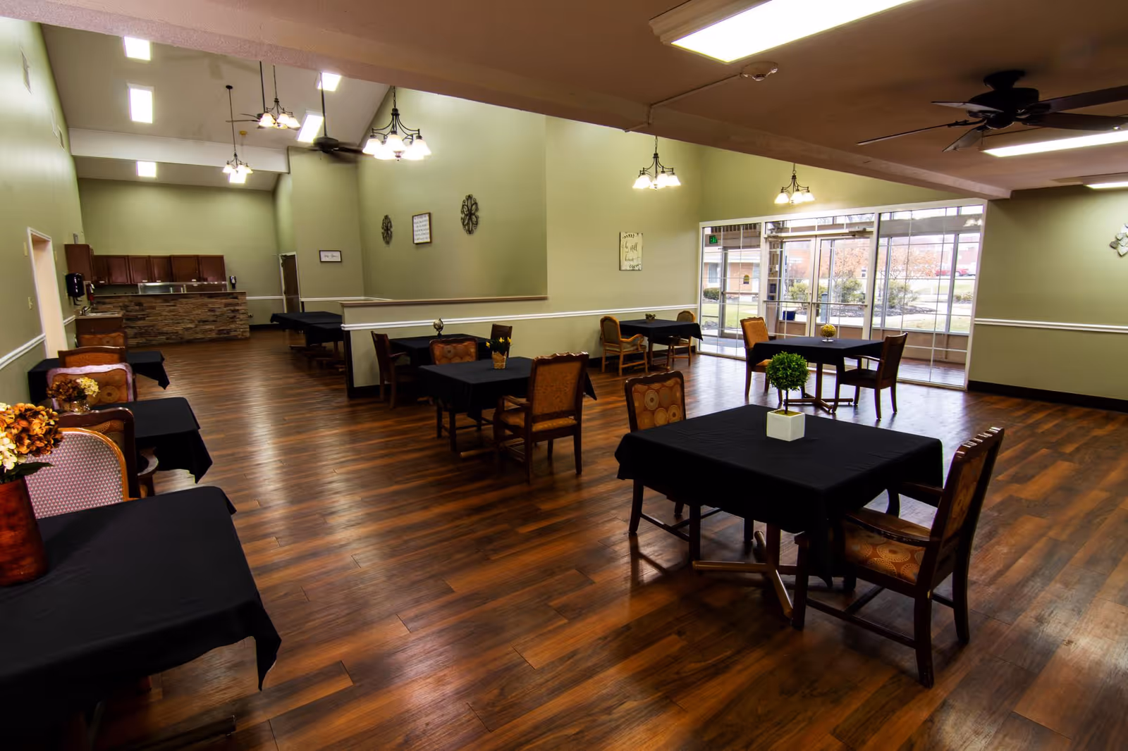 Spacious dining room with wooden floors, several tables covered with black tablecloths, and chairs arranged around them. The room has light green walls, decorative wall hangings, ceiling fans, and hanging light fixtures. Large windows and glass doors allow natural light to enter, showing an outdoor view.