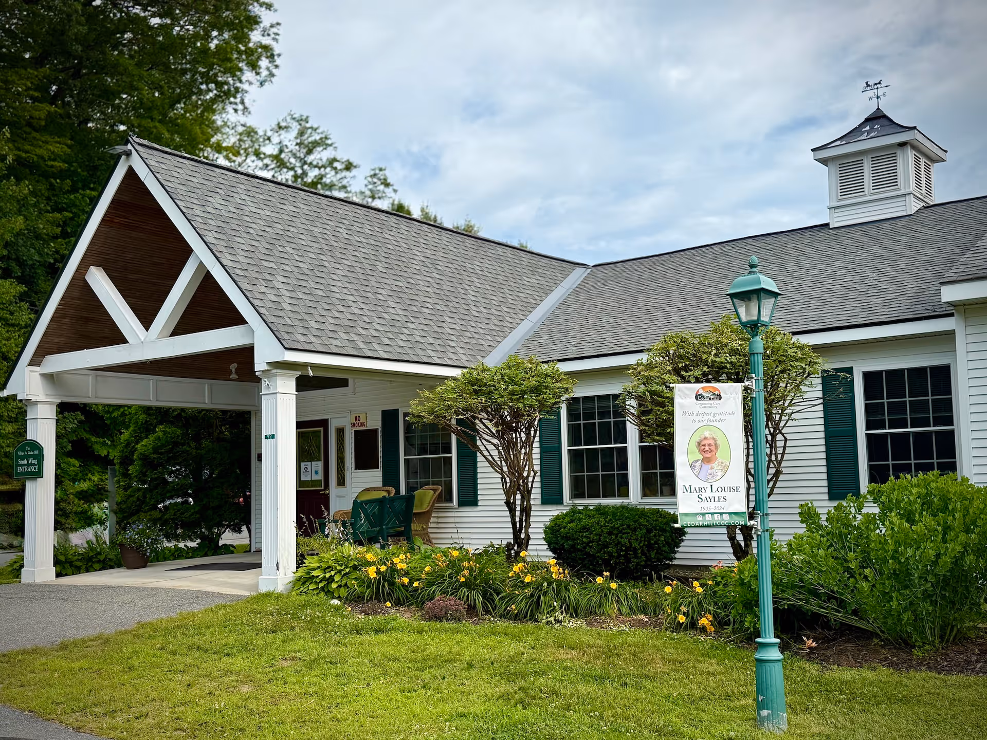 Front entrance of a one-story white community building with a covered portico, green shutters, landscaped flowerbeds and a lamp-post sign featuring a portrait.