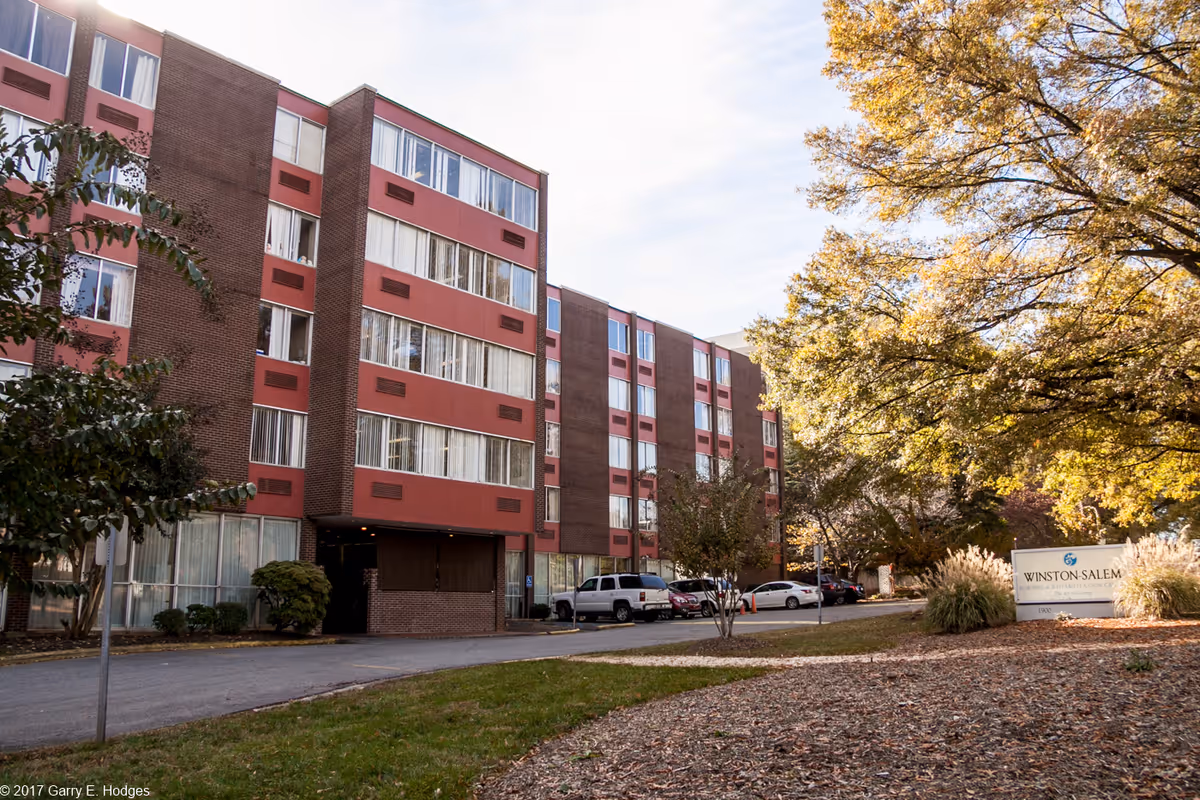 Exterior front of a multi-story brick nursing facility with parked cars and autumn trees on the right.