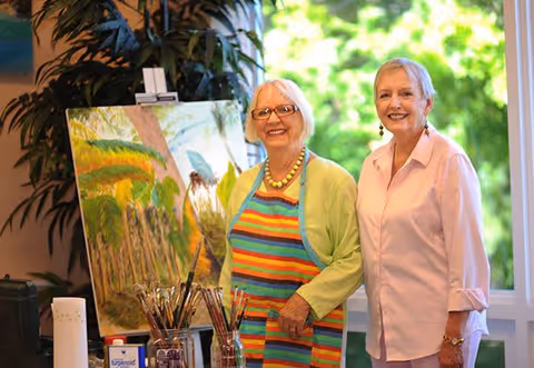 Two elderly women smiling indoors near a window with greenery outside. One woman is wearing a colorful striped apron and glasses, standing next to an easel with a painting of a nature scene. Various paintbrushes and art supplies are on the table in front of them.
