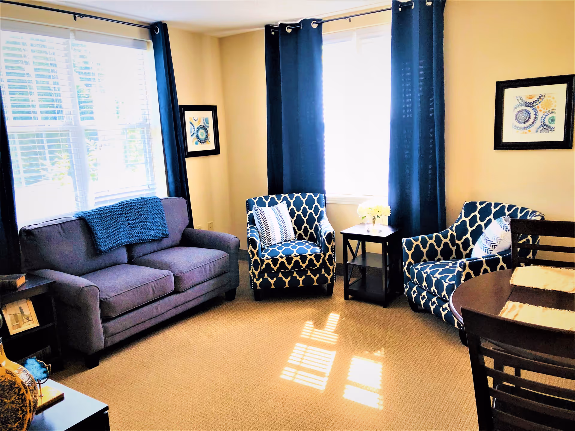 A bright living room with two large windows covered by dark blue curtains. The room features a gray sofa with a blue knitted throw, two patterned blue and white armchairs with decorative pillows, a small dark wooden side table with a vase of flowers, and a dark wooden dining table with chairs. The walls are light-colored and decorated with framed artwork.