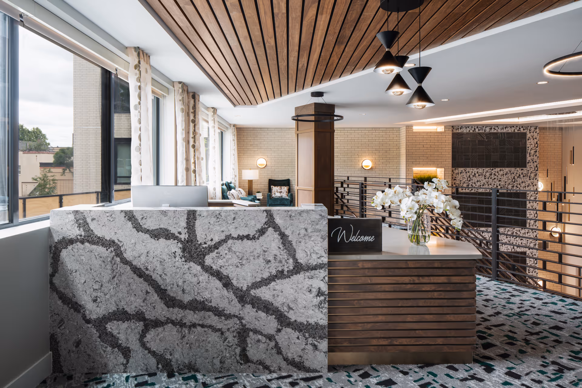 A modern reception area with a marble-patterned front desk and a wooden side counter displaying a 'Welcome' sign and a vase of white orchids. The space features large windows with patterned curtains, a wooden slatted ceiling, contemporary light fixtures, and a seating area with green chairs and a lamp in the background.