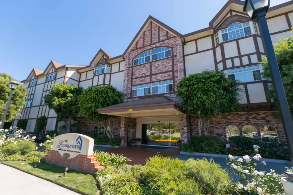 Exterior front view of Lexington Assisted Living facility showing a multi-story building with Tudor-style architecture, brick and stucco facade, large windows, a covered entrance, and landscaped greenery including bushes and trees.