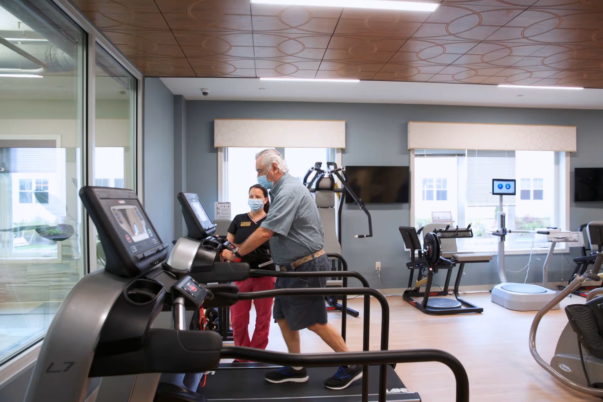 An exercise room where a senior man walks on a treadmill while a staff member assists him, with other gym equipment and windows in the background.