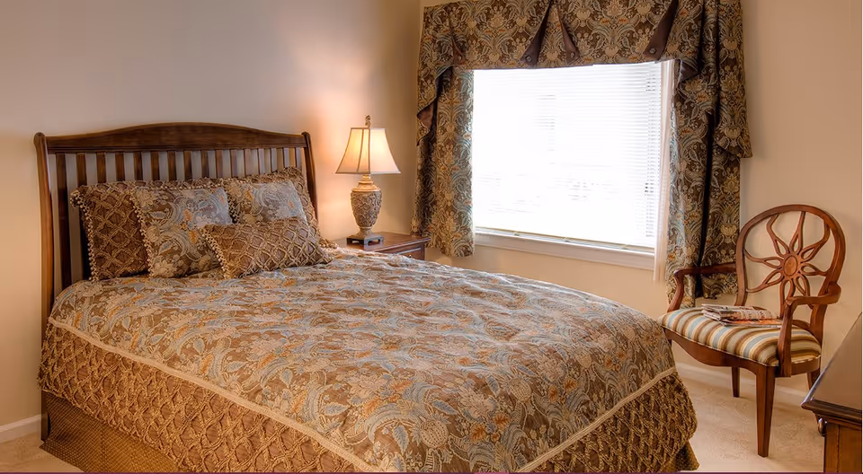 A cozy bedroom with a wooden bed frame and a bed dressed in a patterned comforter and multiple pillows. A bedside table with a decorative lamp is next to the bed. There is a window with patterned curtains and blinds, and a wooden chair with a striped cushion and some magazines on it is placed near the window.