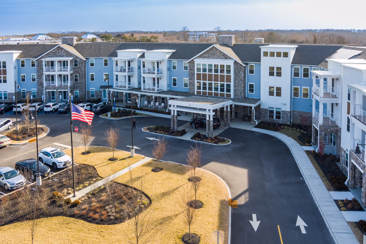 Exterior view of a large senior living facility building with multiple floors, balconies, and a covered entrance. There is a driveway with parked cars, an American flag on a flagpole, landscaped areas with small trees, and a clear sky in the background.