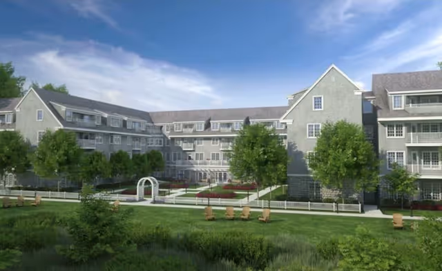 Exterior view of a large senior living facility building with multiple floors, gray siding, and numerous windows. The foreground features a well-maintained green lawn with several wooden chairs arranged in groups, trees, and a white archway leading to a garden area. The sky is clear with some clouds.