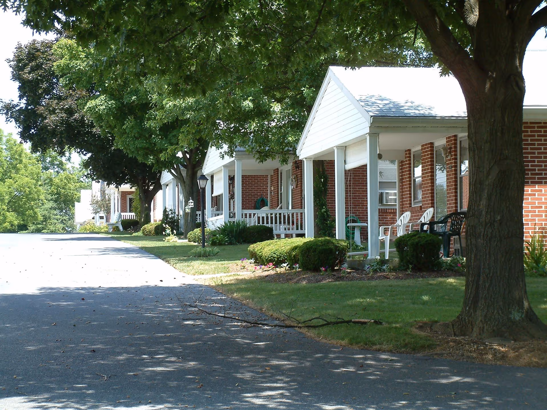 A row of single-story brick residential buildings with white porches and columns, shaded by large trees along a paved driveway. The porches have white and green chairs, and there are well-maintained bushes and flower beds in front of the buildings.