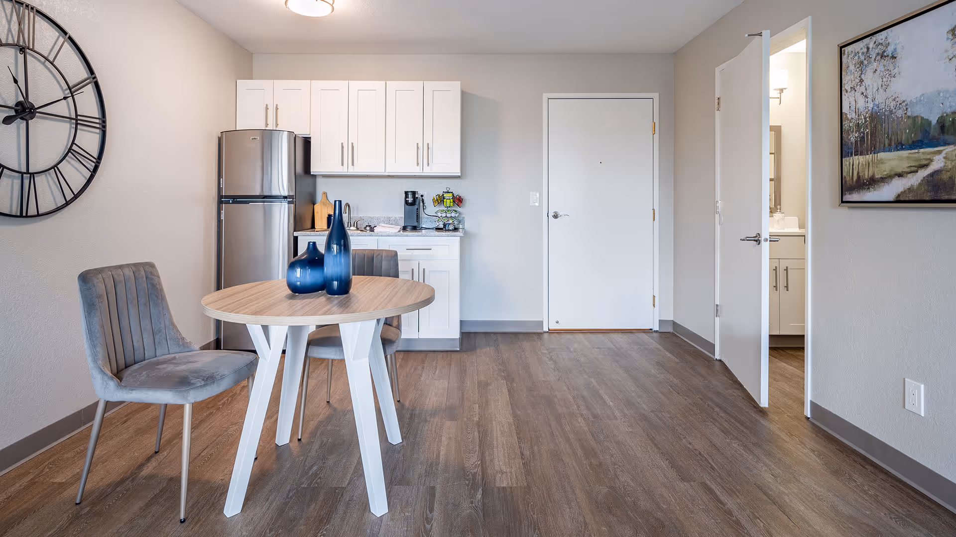 A small dining area with a round wooden table and two gray upholstered chairs. On the table are three decorative blue vases. Behind the table is a compact kitchen with white cabinets, a stainless steel refrigerator, and a coffee maker on the counter. To the right, there is a closed white door and an open door leading to a bathroom with a sink and mirror. A large wall clock is mounted on the left wall, and a framed landscape painting hangs on the right wall. The floor is covered with wood-style flooring.