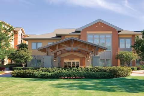 Front exterior view of a senior living community building with a large covered entrance, well-maintained green lawn, shrubs, and trees under a partly cloudy sky.