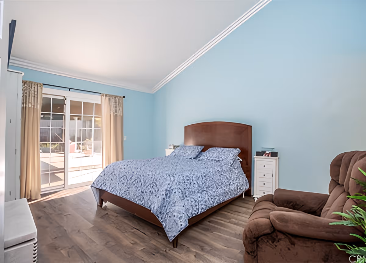 Bright bedroom with a bed against a blue wall, sliding glass doors, and a brown recliner.