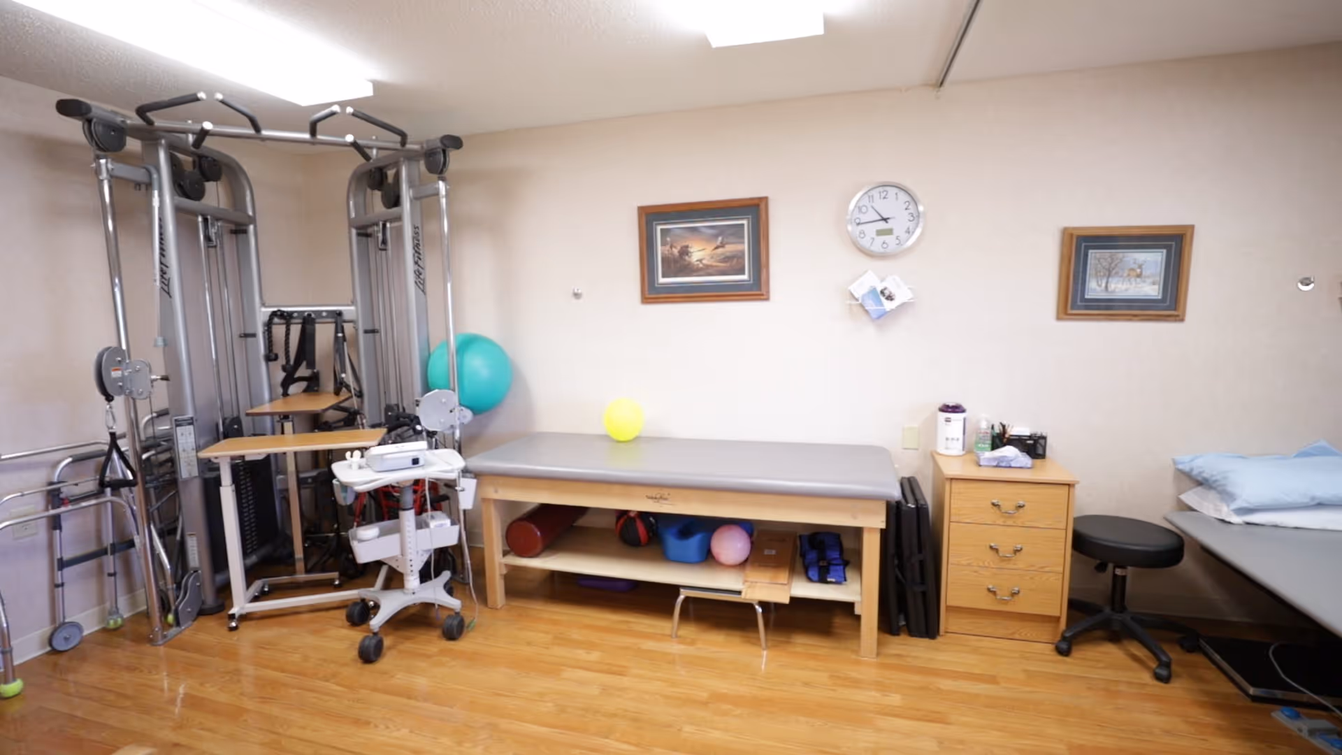 A physical therapy or rehabilitation room with exercise equipment, therapy tables, a walker, exercise balls, and a rolling stool. The room has wooden flooring, beige walls with two framed pictures, and a clock on the wall.