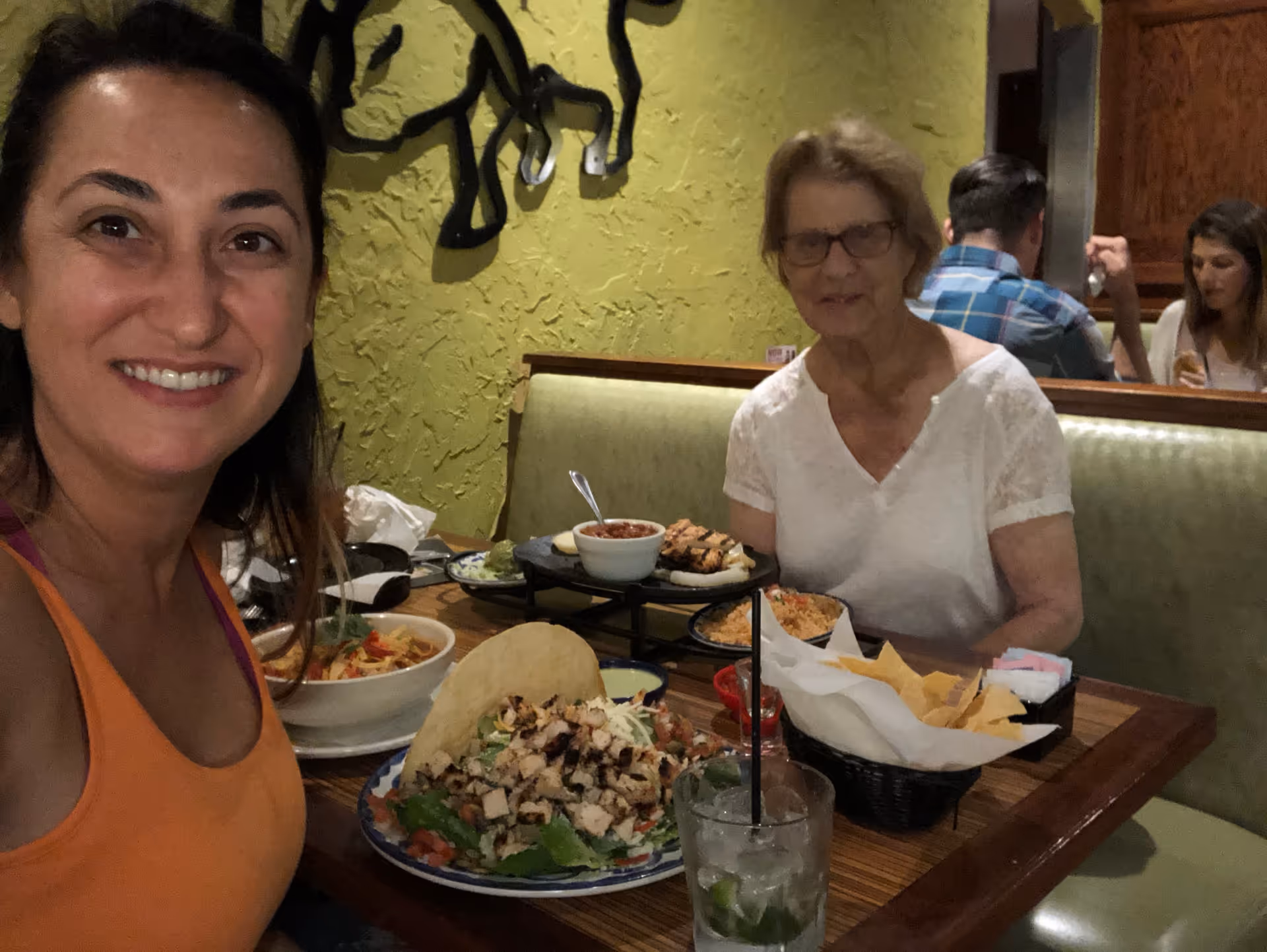 Two women sit at a restaurant booth smiling at the camera with plates of tacos, chips, and drinks on the table.