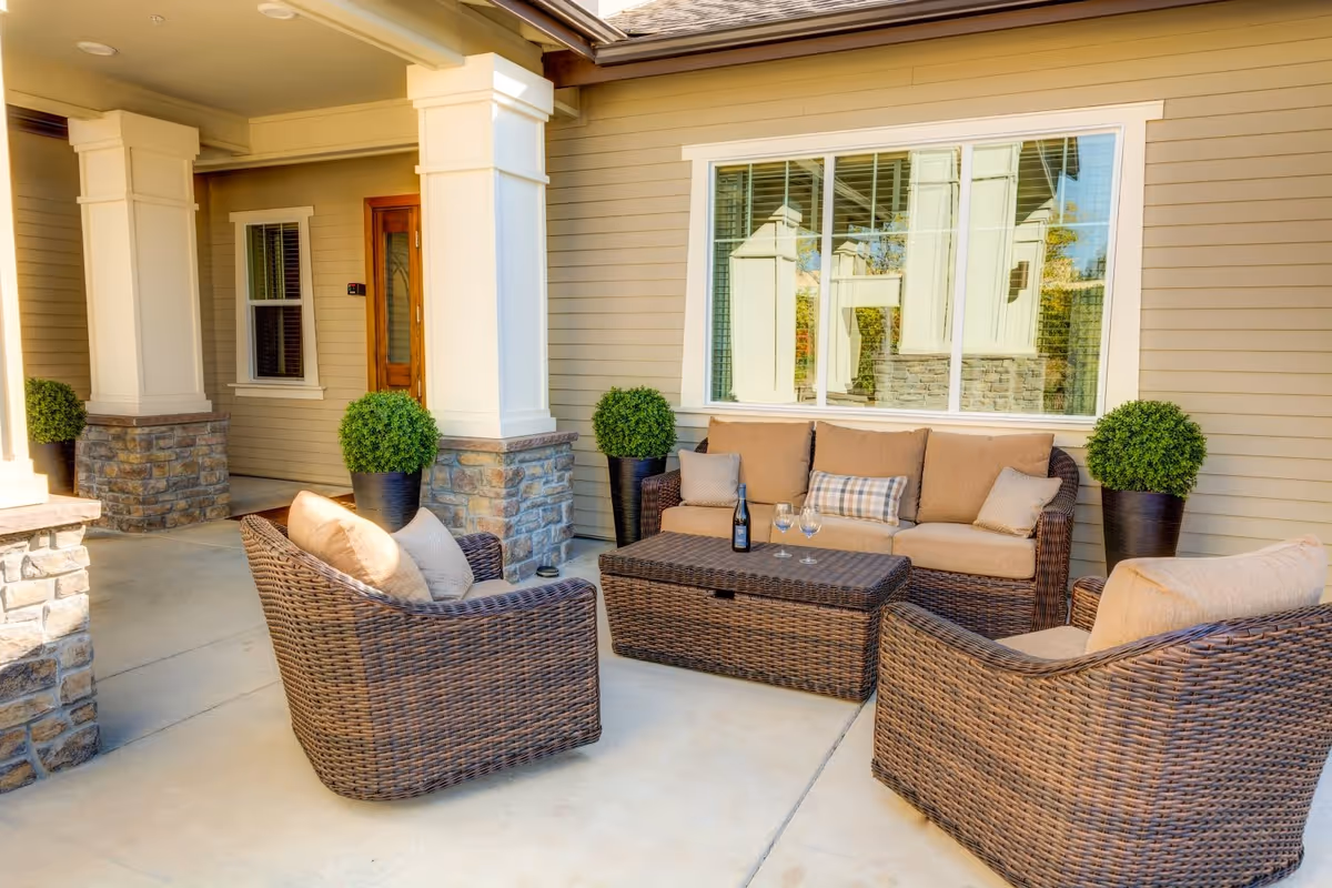 Outdoor patio area with wicker furniture including a sofa and two armchairs with beige cushions, a wicker coffee table with a bottle and three wine glasses on it, potted green plants, and a beige house exterior with white trim and large windows.