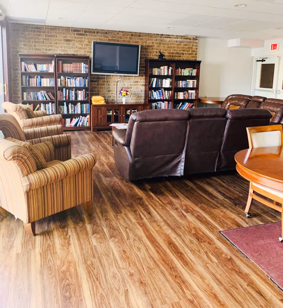 A cozy living room area with wooden flooring, two striped armchairs, a brown leather sofa, a round wooden table with chairs, and bookshelves filled with books against a brick wall. A flat-screen TV is mounted on the brick wall above a wooden cabinet with a vase of flowers on top.