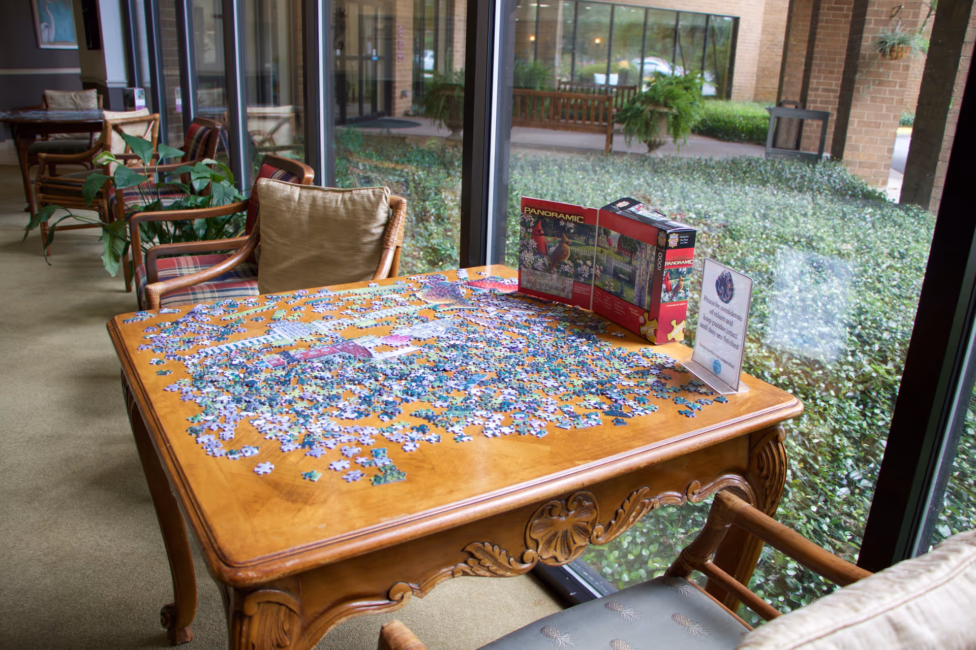 A wooden table with an incomplete jigsaw puzzle spread out on it, located next to large windows overlooking greenery outside. The puzzle box and a sign asking to keep puzzle pieces until finished are also on the table. Several chairs and plants are arranged along the windowed hallway.