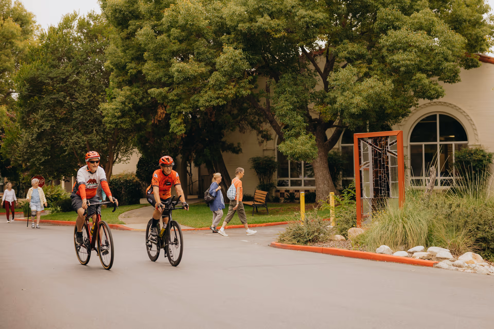 Two bicyclists ride on a driveway in front of a building while pedestrians walk among trees and landscaping.