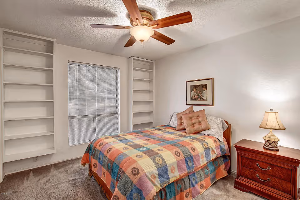 A bedroom with a bed covered in a colorful patchwork quilt, two decorative pillows, a wooden nightstand with a lamp, built-in white shelves on either side of a window with blinds, and a ceiling fan with a light fixture.