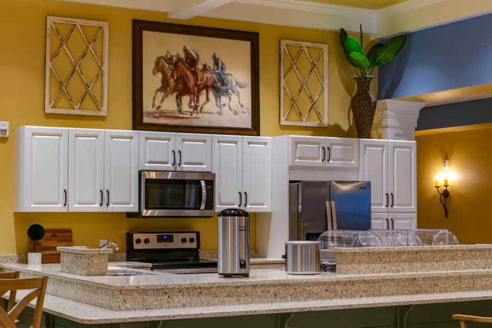 A modern kitchen area with white cabinets, a stainless steel microwave and stove, and a stainless steel refrigerator. The countertop is made of speckled granite and has a coffee urn and a covered food display. Above the cabinets, there is a framed painting of horse riders and decorative wall hangings. A large vase with green plants is placed on top of the cabinets. The walls are painted yellow and blue, and there is a wall sconce light fixture on the right side.
