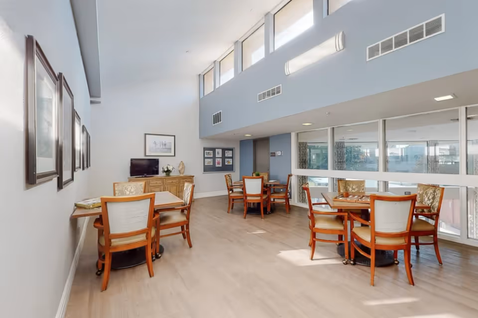 A bright and spacious common area with several wooden tables and chairs arranged for seating. The room features large windows allowing natural light to fill the space, light-colored walls, and framed artwork on the walls. A small TV and decorative items are placed on a wooden cabinet against one wall.