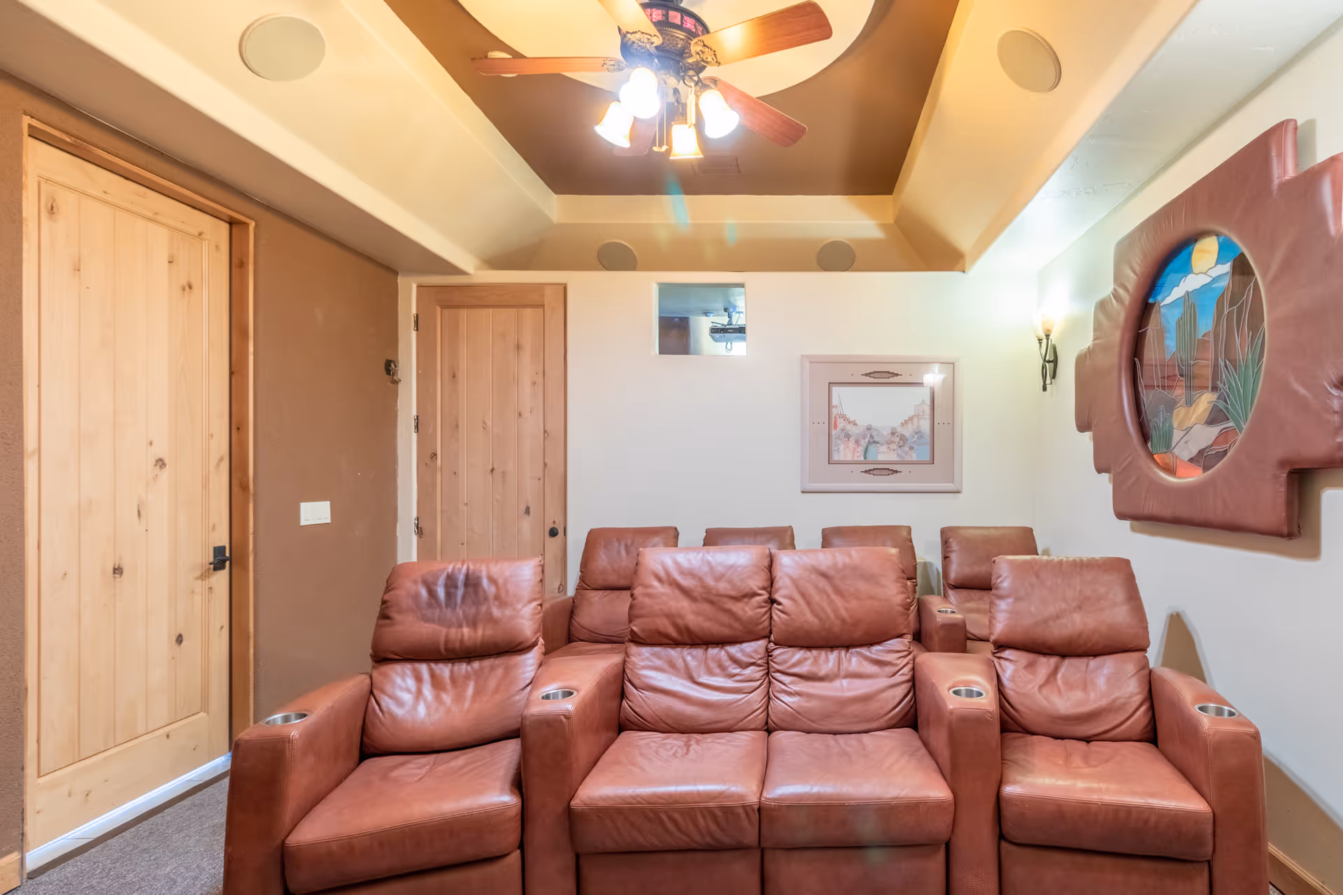 A cozy home theater room with two rows of brown leather recliner chairs, each equipped with cup holders. The room features a ceiling fan with lights, a small window on the back wall, a framed picture, and a decorative wall piece with a desert scene. The walls are painted in warm tones, and there are two wooden doors on the left side.
