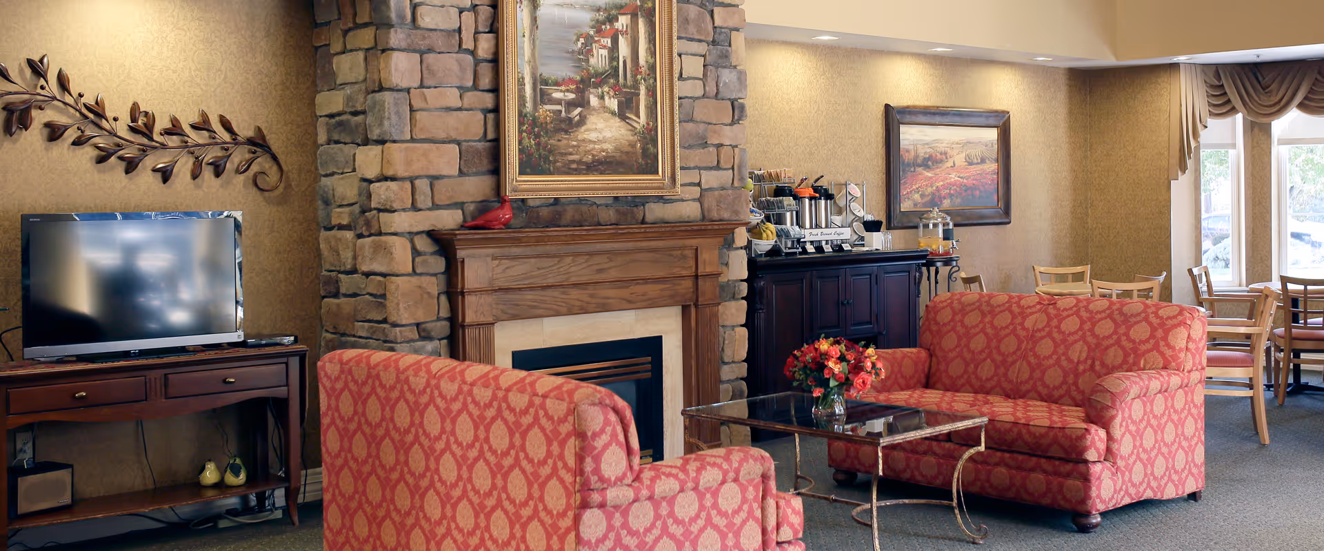 A cozy senior living room featuring two red patterned armchairs and a matching loveseat arranged around a glass coffee table with a flower vase. A stone fireplace with a wooden mantle and a framed painting above it is centered in the room. To the left, there is a wooden TV stand with a flat-screen television and decorative items. In the background, a dining area with wooden tables and chairs is visible near large windows with draped curtains.