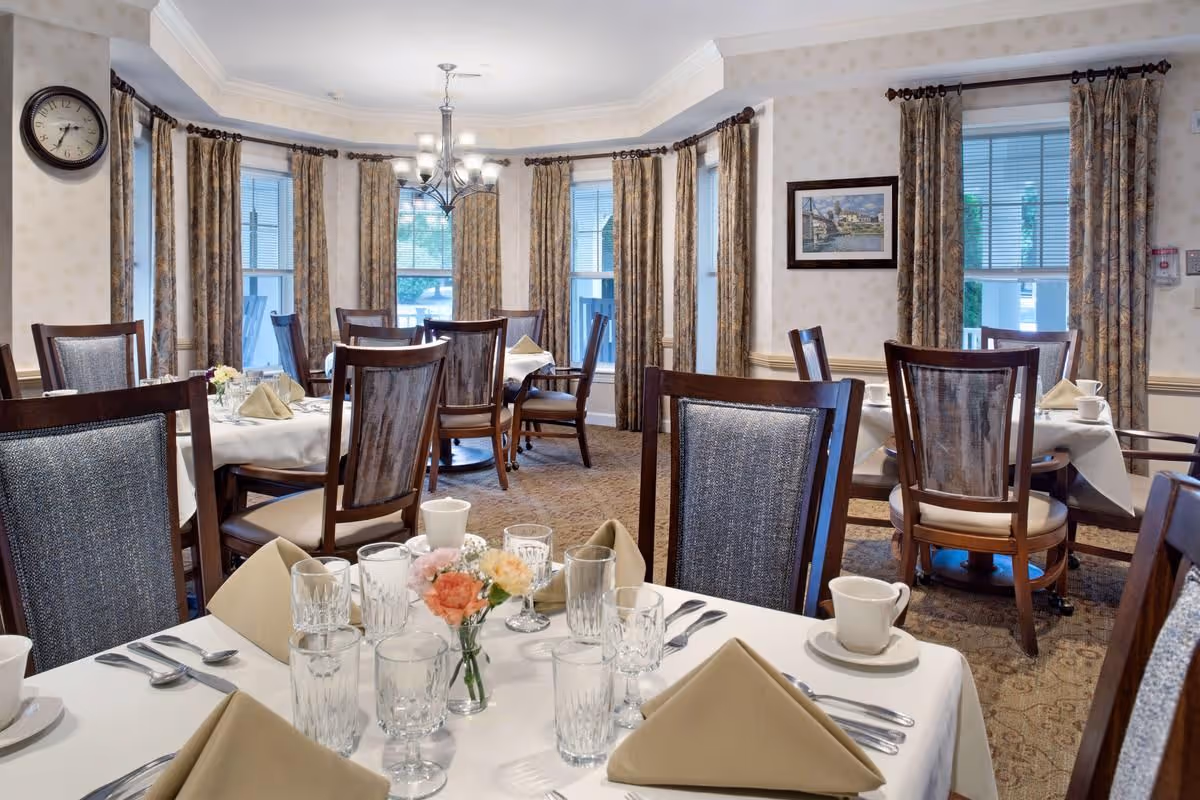 Well-appointed dining room with tables set with glassware, napkins, and floral centerpieces, surrounded by wooden chairs and curtained windows.