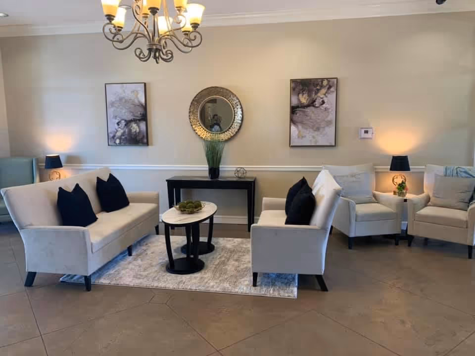 A cozy living room area with two beige sofas facing each other, each with black throw pillows, and two beige armchairs with cushions. A small oval coffee table with decorative items sits on a light-colored rug between the sofas. Behind the seating area is a black console table with a round decorative mirror and a plant. The walls are beige with two abstract paintings and two table lamps providing warm lighting. A chandelier hangs from the ceiling.