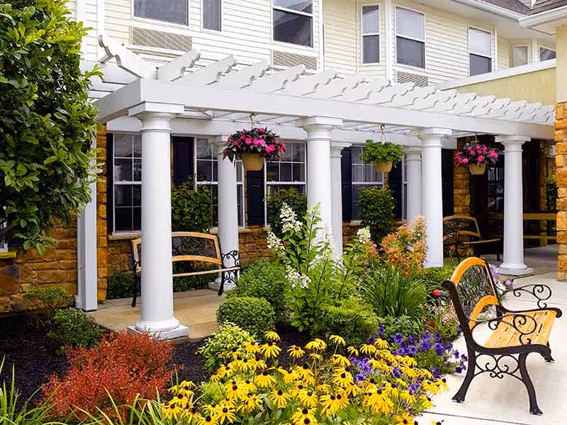 Sunny outdoor courtyard with a white pergola supported by columns, benches, hanging flower baskets and a colorful planted garden in front of a building.