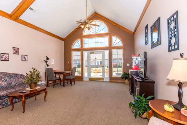 A bright communal living room with a vaulted ceiling, sofa and chairs, a TV console and side tables, and large arched windows and doors leading outside.