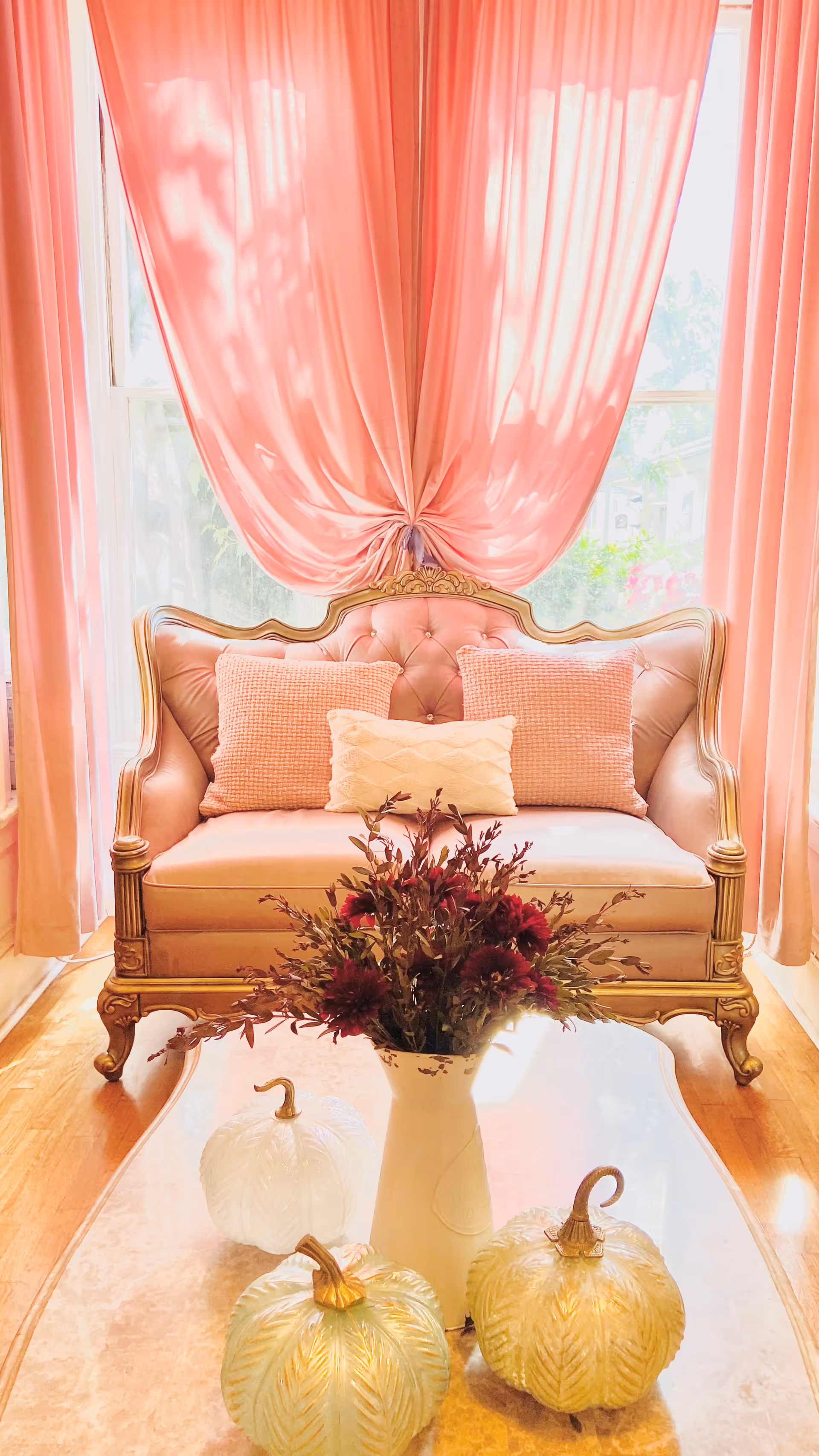 A cozy living room area with a vintage-style pink tufted loveseat adorned with three pillows in shades of pink and cream. Behind the loveseat, sheer pink curtains are draped over a large window letting in natural light. In front of the loveseat, there is a glass coffee table with a white vase holding dark red flowers and three decorative glass pumpkins in white and gold tones.