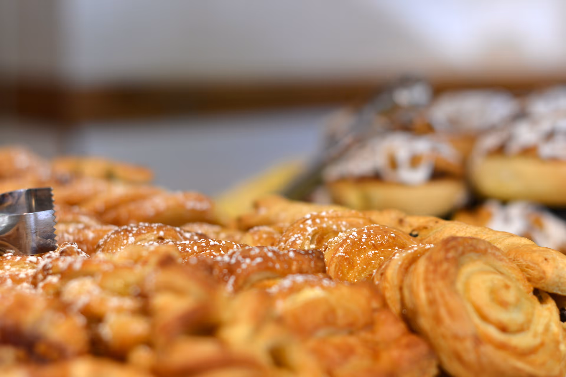 Close-up view of a variety of freshly baked pastries, including croissants and cinnamon rolls, some sprinkled with powdered sugar, arranged on a tray with tongs visible on the left side.