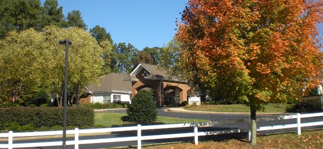 Front exterior of a brick senior living building with a driveway, white fence, and autumn trees.