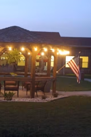 Outdoor wooden gazebo with hanging plants and string lights illuminated at dusk, with an American flag mounted on one side. The gazebo is surrounded by grass and a building with lit windows is visible in the background.