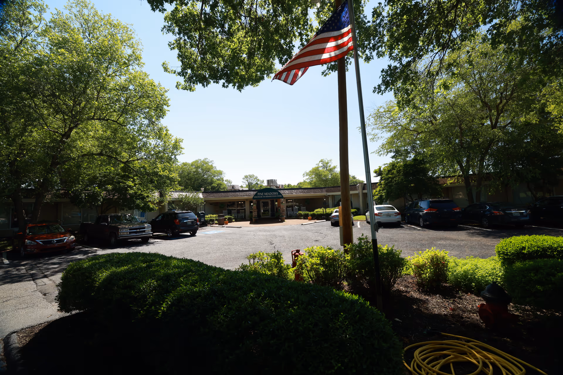 View of the front exterior of Pine Meadows Healthcare Center with a parking lot, several cars, an American flag on a pole, and surrounding trees and bushes under a clear sky.