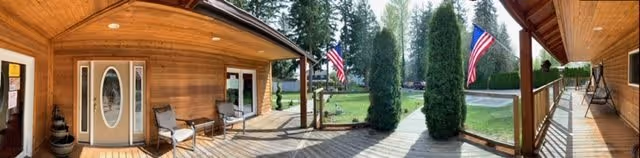 Covered wooden front porch with chairs, an oval-glass entry door, two American flags, tall columnar shrubs, and a view of the lawn and driveway.