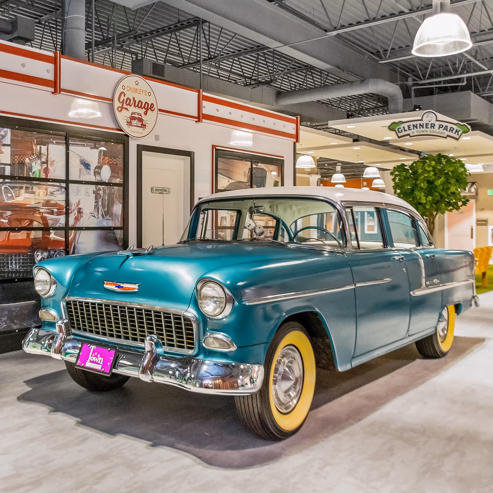 A vintage teal and white Chevrolet car with yellow whitewall tires displayed indoors in front of a garage-themed wall with a sign reading 'Chumley's Garage'. The setting appears to be an indoor common area with overhead lighting and a sign for 'Glenner Park Town Square' visible in the background.