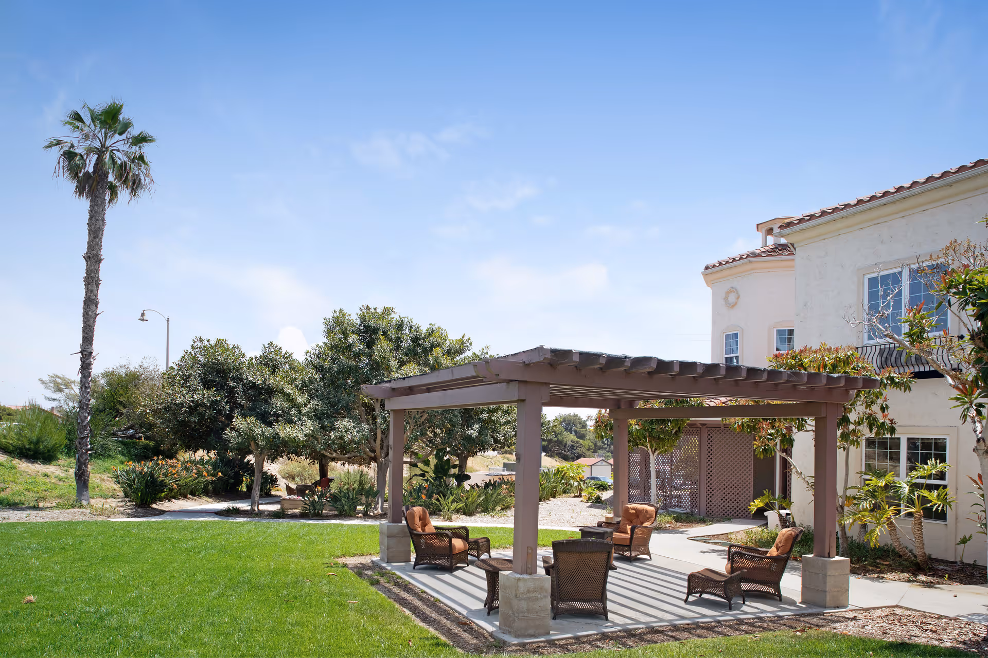 Outdoor seating area with a wooden pergola providing shade over several cushioned wicker chairs and a small table, surrounded by green grass, trees, and shrubs, next to a light-colored building under a clear blue sky.